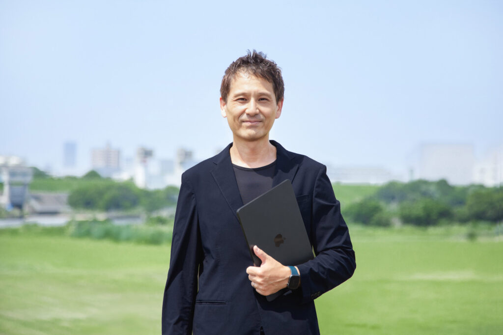 河川敷でノートパソコンを持って微笑んでいるジャケットを着た男性